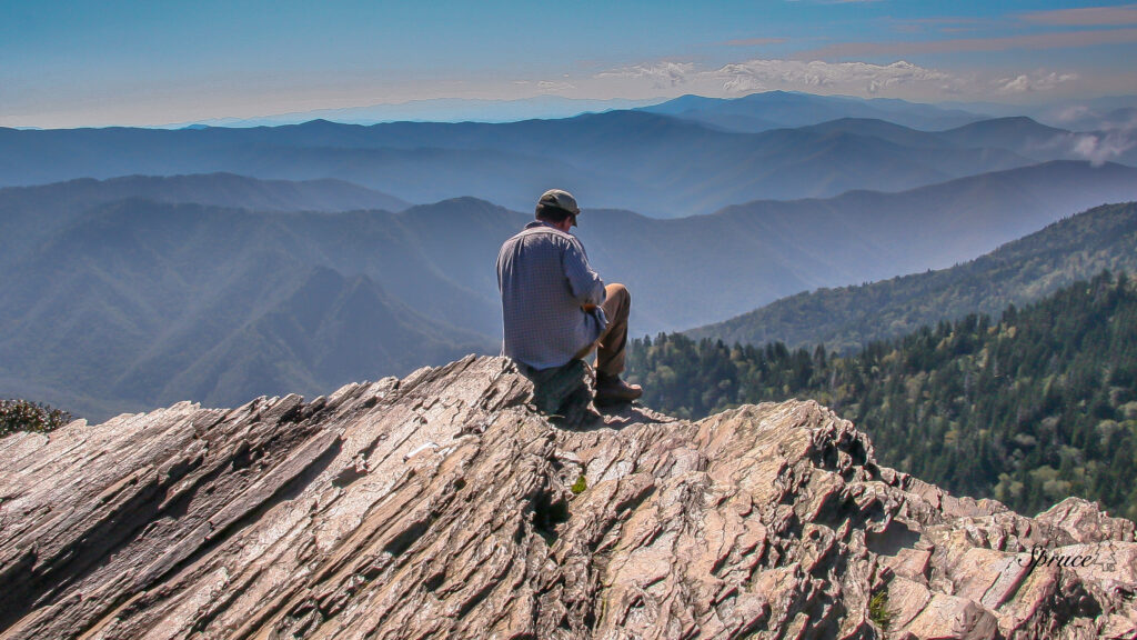 Person sitting on a rocky outcrop in the center of the image with rows of mountains in the background.