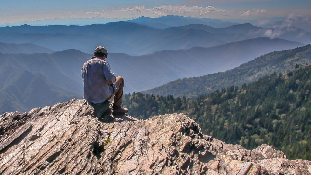 Person sitting on a rocky out-crop to the left of the image with rows of mountains in the background.